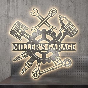 May include: A dark metal wall sign featuring a central gear with the text MILLER'S GARAGE. The design includes a piston, a wrench, an impact driver, two spark plugs, and a ratchet arranged around the gear, all illuminated by a warm backlighting.