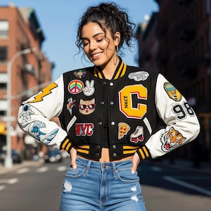 Puede incluir: Una chaqueta universitaria en blanco y negro con ribetes acanalados a rayas amarillas y numerosos parches coloridos, incluyendo una letra C amarilla grande, un gato con gafas de sol, un signo de paz, un tigre, una porción de pizza y el texto NYC.