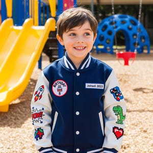 May include: A navy blue and cream varsity jacket featuring white striped ribbed trim, a round birthday patch with a cake and the number 8, a rectangular patch reading Joined: 2023, and various colorful patches including a rocket, dinosaur, and heart.