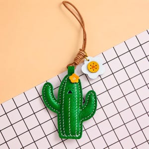 Puede incluir: Un llavero con forma de cactus verde con costuras blancas, un pequeño detalle de flor naranja y un cordón de cuero marrón. Unido al cordón hay un dije con forma de flor blanca con un centro amarillo. El cactus tiene un rostro triangular negro.
