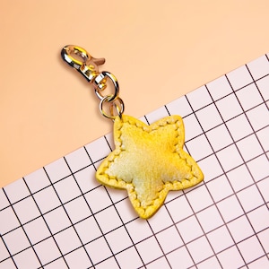 May include: A yellow star-shaped keyring featuring a textured surface with visible stitching along the edges. The star is attached to a gold-toned metal lobster clasp by a small circular jump ring, providing a secure way to attach the item.