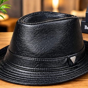 Pode incluir: Um chapéu fedora preto com um acabamento texturizado semelhante a couro. O chapéu apresenta uma faixa preta a condizer à volta da copa e um pequeno emblema metálico prateado de forma triangular na lateral da faixa com um design subtil.