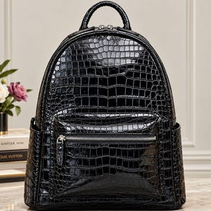 May include: A black backpack with a glossy, embossed crocodile skin texture. The bag features a rounded top handle, a main zippered compartment, and a smaller front zippered pocket with a silver-toned metal zipper pull and hardware.