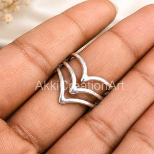 Puede incluir: Un anillo de tono plateado con un diseño de triple chevrón que presenta tres bandas apiladas en forma de V que convergen en los lados. El metal tiene un acabado liso y pulido, y el anillo está colocado en un dedo, mostrando la estructura.