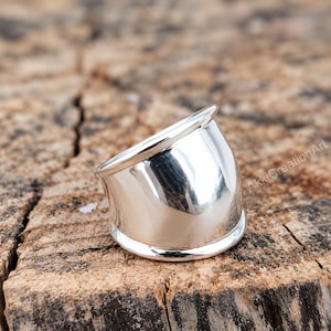 May include: A wide, polished silver ring with a smooth, reflective surface and a curved, tapered silhouette. The band features a rounded edge at the top and bottom, creating a sleek, solid metallic appearance with the text AkkiCreationArt visible.