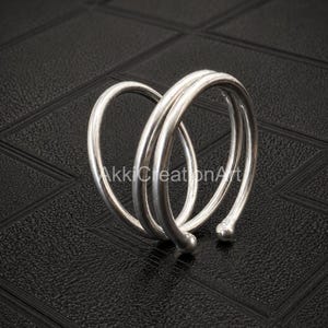 May include: A silver-coloured metal ring featuring a bypass spiral design with two overlapping bands. Each end of the wire terminates in a small, smooth spherical bead. The surface of the metal has a polished, reflective finish throughout the piece.