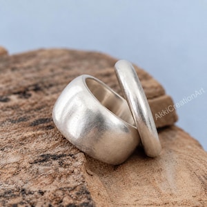 Puede incluir: Dos anillos de tono plateado con un acabado liso y mate. Un anillo presenta una banda ancha y gruesa, mientras que el otro es una banda más delgada y redondeada. El texto AkkiCreationArt es visible en el lado derecho de la imagen.