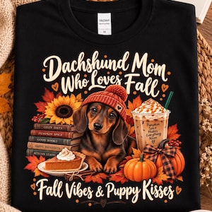 Peut inclure: Un t-shirt noir avec un motif de teckel portant un bonnet en tricot rouge, entouré de feuilles d'automne, d'un tournesol, de livres, d'une part de tarte à la citrouille, d'une citrouille et d'un café avec le texte "Dachshund Mom Who Loves Fall" et "Fall Vibes & Puppy Kisses".