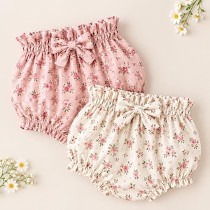 May include: Two pairs of bloomers featuring a ruffled elastic waistband and a decorative bow at the center. One pair is light pink with a small floral pattern, and the other is off-white with a matching pink and green floral print design.