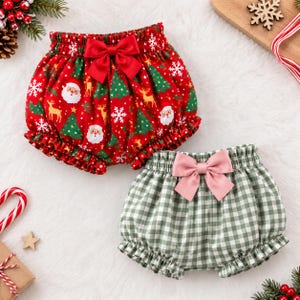 May include: Two pairs of ruffled bloomers. One pair is red with a Christmas-themed print featuring Santa Claus, reindeer, pine trees, and white snowflakes, accented with a red bow. The other pair is green and white gingham with a pink bow.