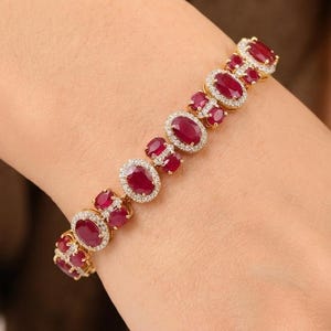 Puede incluir: Una pulsera de oro con una serie de rubíes de color rojo intenso en talla ovalada, cada uno rodeado por un halo de pequeños diamantes blancos. La pulsera está construida con engastes de rubí alternos conectados por grupos de rubíes y diamantes.