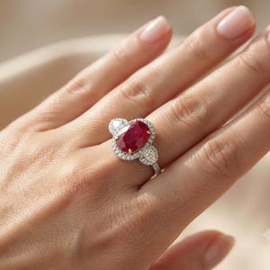 Può includere: Un anello con una grande pietra centrale di rubino rosso intenso a taglio ovale, circondata da un alone di piccoli diamanti bianchi rotondi e chiari. Due diamanti a taglio pera sono incastonati ai lati del rubino su una sottile fascia.