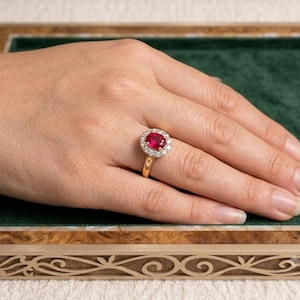 Op de afbeelding: Een gouden ring met een centrale ovaal geslepen levendige rode edelsteen, omringd door een halo van kleine ronde witte diamanten. De ring wordt om een vinger gedragen en toont de gepolijste gouden band en het contrast tussen de stenen.