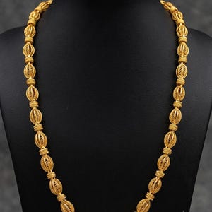 Puede incluir: Un collar de cadena dorada que presenta una serie de cuentas filigranas ovaladas y alargadas. Cada cuenta está elaborada con un intrincado trabajo de alambre y separada por pequeños anillos dorados texturizados, creando un patrón uniforme.