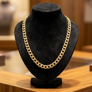 Puede incluir: Un collar de cadena tipo barbada en tono dorado con eslabones ovalados entrelazados y un acabado texturizado brillante. El collar se exhibe sobre un busto cubierto de terciopelo negro, mostrando el tamaño uniforme de los eslabones metálicos.