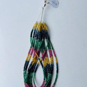 Op de afbeelding: Een meerstrengs ketting bestaande uit kleine, gefacetteerde edelsteenkralen in levendige tinten geel, blauw, groen en roze. De strengen zijn bovenaan samengebonden met een wit label met handgeschreven tekst, vastgezet met dun metaaldraad.
