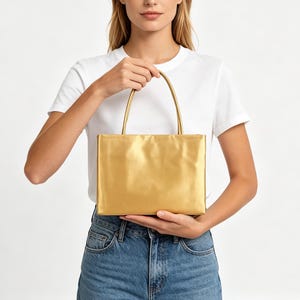 May include: A rectangular handbag with a metallic gold finish and a smooth, solid surface. The bag features two thin, matching gold handles attached to the top edge, providing a structured and clean appearance for this compact accessory.