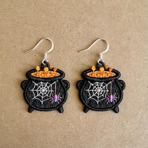 May include: A pair of embroidered earrings featuring black cauldrons with orange bubbling liquid at the top. Each cauldron displays a white spiderweb design and a small purple spider hanging from a thread, attached to silver-toned metal hooks.