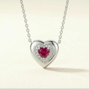 May include: A silver heart-shaped pendant necklace featuring a central heart-cut red gemstone surrounded by a halo of small white stones, suspended from a delicate silver chain with a polished metallic finish and a smooth, rounded frame.