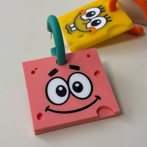 Puede incluir: Un par de pendientes cuadrados con caras de personajes de dibujos animados. Un pendiente es rosa con un aro verde azulado y el otro es amarillo con un aro naranja. Ambas piezas tienen una textura porosa con ojos grandes y bocas sonrientes.