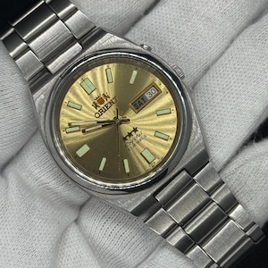 May include: A silver-toned stainless steel wristwatch with a round gold-coloured dial. The watch features a metal link bracelet, luminous hour markers, and a day-date window at the three o'clock position. The dial displays the Orient logo and text.