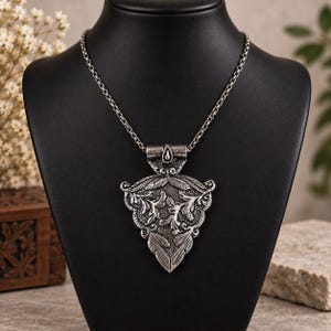 May include: A silver-toned metal pendant necklace featuring an ornate, triangular-shaped charm with intricate embossed leaf and scroll patterns. The pendant hangs from a sturdy, textured silver-toned chain, showcasing a dark, oxidized finish.