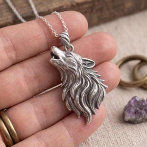 Puede incluir: Un collar de tono plateado con un colgante detallado en forma de cabeza de lobo aullando. El colgante tiene un acabado texturizado y oxidado que resalta los detalles del pelaje y la boca abierta del lobo. Cuelga de una cadena plateada fina.