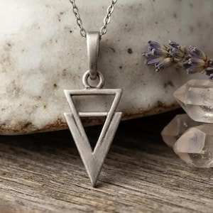 May include: A silver-toned metal necklace featuring a pendant shaped like two stacked triangles with a horizontal bar across the centre. The pendant hangs from a delicate silver-toned chain, showcasing a matte finish and geometric design elements.