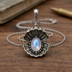 May include: A silver pendant shaped like an open seashell with a central oval moonstone cabochon. The pendant features detailed ridges and a polished finish, suspended from a delicate silver chain. The moonstone displays a blue iridescent glow.