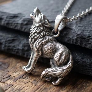 May include: A silver-toned metal pendant shaped like a wolf howling with its head tilted upwards. The pendant features detailed fur texture and is attached to a silver-toned metal chain link necklace. The metal has a polished and oxidised finish.