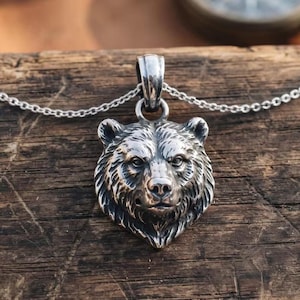 Puede incluir: Un colgante de metal plateado con forma de cabeza de oso detallada, que presenta patrones de pelaje texturizados y rasgos faciales definidos. El colgante está unido a una cadena de metal plateado fina y a juego que se extiende horizontalmente.
