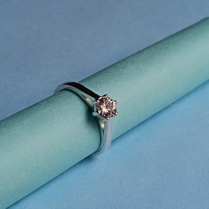 Könnte beinhalten: Ein silberfarbener Ring mit einem einzelnen, rund geschliffenen, klaren Edelstein in einer sechs-krappigen Fassung. Die Ringschiene hat eine glatte, polierte Oberfläche und verjüngt sich zum Stein hin, was für ein klares Design sorgt.