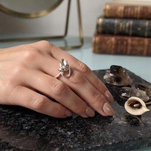 Könnte beinhalten: Ein silberner Ring mit einem skulpturalen, organischen Design, das an eine geknotete oder verdrehte Form erinnert. Das Metall hat eine polierte, reflektierende Oberfläche und das Band ist schmal und umschließt den Finger in einer fließenden Form.