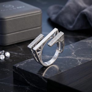 Könnte beinhalten: Ein silberfarbener offener Ring mit zwei parallelen Stäben, die mit kleinen klaren Steinen besetzt sind. Der Ring hat ein strukturelles, geometrisches Design mit durchbrochenen Dreiecksmustern an den Seiten, was ihm eine architektonische Optik verleiht.
