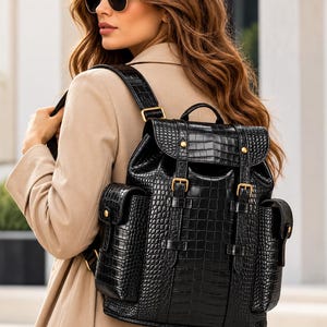 May include: A black backpack with a crocodile-embossed texture, featuring two side pockets, a top flap with two buckle straps, and gold-toned metal hardware. The bag has adjustable shoulder straps and a small top handle for carrying.