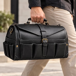 Pode incluir: Uma bolsa de ferramentas preta estruturada com uma alça superior, um fecho central com fivela e vários bolsos exteriores na parte frontal e nas laterais. A bolsa apresenta cantos reforçados e ferragens em metal com tom de latão envelhecido.