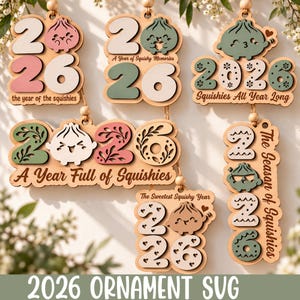 以下が含まれることがあります： 「the year of the squishies」、「A Year of Squishy Memories」、「Squishies All Year Long」、「A Year Full of Squishies」、「The Season of Squishies」というテキストと様々な餃子キャラクターが描かれた5つの木製2026年オーナメントのコレクション。