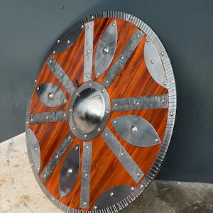 Pode incluir: Um escudo circular de madeira com um umbo central de metal polido, tiras metálicas radiantes e detalhes em forma de folha. O escudo possui uma borda de metal texturizada e é fixado com rebites visíveis sobre a superfície de madeira.