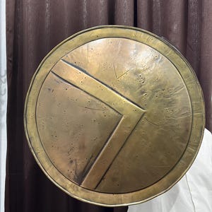 May include: A circular brass shield featuring a prominent raised Lambda symbol in the centre. The surface of the shield displays a weathered, textured finish with various small indentations, scratches, and dark patina spots across the metal.