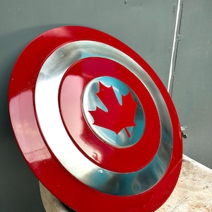 May include: A circular shield featuring a glossy red outer ring, a metallic silver middle ring, and a central silver disc adorned with a bold red maple leaf emblem. The design is smooth and reflective, showcasing a clean, polished finish throughout.