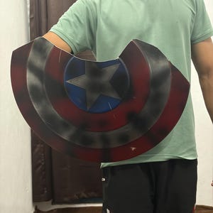 May include: A circular shield featuring a central blue circle with a silver star, surrounded by concentric rings of red and black. The shield has a weathered, distressed finish with dark shading and a notched cutout along the top edge.
