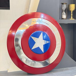 May include: A circular shield featuring a red outer ring, a silver middle ring, and a blue center circle with a silver star. The silver ring is engraved with the text To a Super Dad - Love E and F. The shield has a smooth, reflective finish.