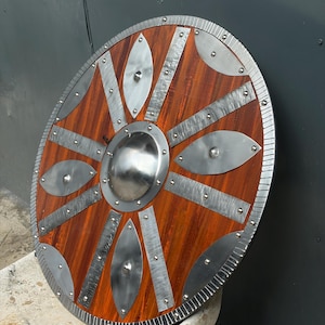 May include: A circular wooden shield featuring a reddish-brown wood grain finish. The shield is reinforced with a central steel boss, radiating steel strips, and leaf-shaped steel plates, all secured with visible rivets along the textured rim.