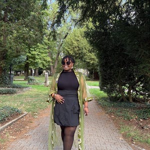 May include: A black mock neck top and a black skirt paired with a sheer olive green duster featuring a white and yellow floral pattern along the front edges, worn with black tights, black high-heeled shoes, and dark-tinted sunglasses.