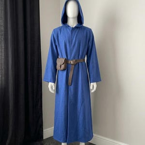 Pode incluir: Uma túnica azul com capuz que chega ao chão, com mangas compridas e gola alta. A peça é ajustada na cintura com um cinto de couro castanho que apresenta uma pequena bolsa de couro castanho presa no lado direito, sobre o tecido azul.