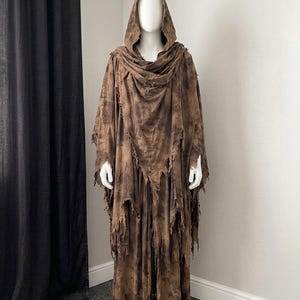May include: A full-length brown hooded robe featuring a distressed, tattered appearance with uneven, frayed edges and a mottled, dark-toned fabric texture. The garment includes a deep hood and long, flowing sleeves that drape over the mannequin.
