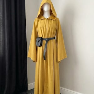 Pode incluir: Uma túnica com capuz de cor amarelo-mostarda que chega ao chão, com mangas compridas e largas. A peça é ajustada na cintura com um cinto preto que possui uma pequena bolsa preta acoplada. A túnica tem uma abertura frontal e um capuz.