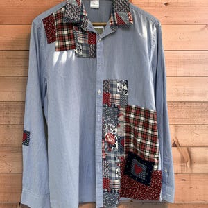 Può includere: Una camicia a maniche lunghe a righe blu e bianche con dettagli patchwork. Il patchwork include tessuti a fantasia rossi, bianchi e blu, tra cui quadri e disegni floreali. La camicia è abbottonata e appesa a una gruccia di legno.