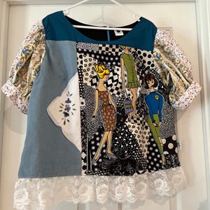 Könnte beinhalten: Ein Patchwork-Oberteil mit verschiedenen Stoffen, darunter Jeansstoff, Blumenmuster und ein schwarz-weißes Muster mit Illustrationen von Frauen. Das Oberteil hat einen tealfarbenen Ausschnitt und Ärmel mit floralen Manschetten sowie eine weiße Spitzenborte am Saum.