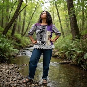 Può includere: Un top patchwork con un mix di motivi bianchi e neri, accenti viola e rifiniture in pizzo. Il top ha maniche a tre quarti ed è abbinato a jeans blu. La persona è in piedi in una foresta.